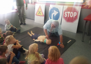 Na materacu leży manekin dziecka. Bada go stetoskopem mężczyzna przebrany za lekarza. Wokół materaca siedzą dzieci.